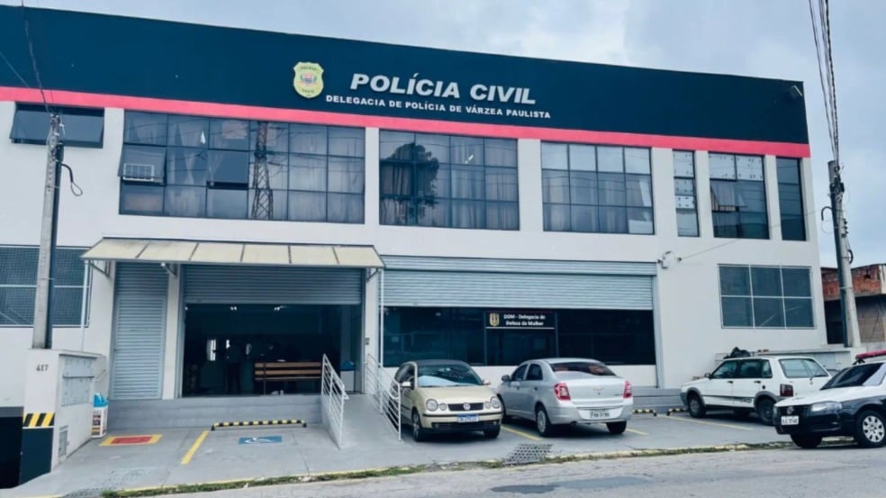 Fachada da Delegacia de Polícia de Várzea Paulista, com carros estacionados em frente e o letreiro da Polícia Civil visível no topo do prédio.