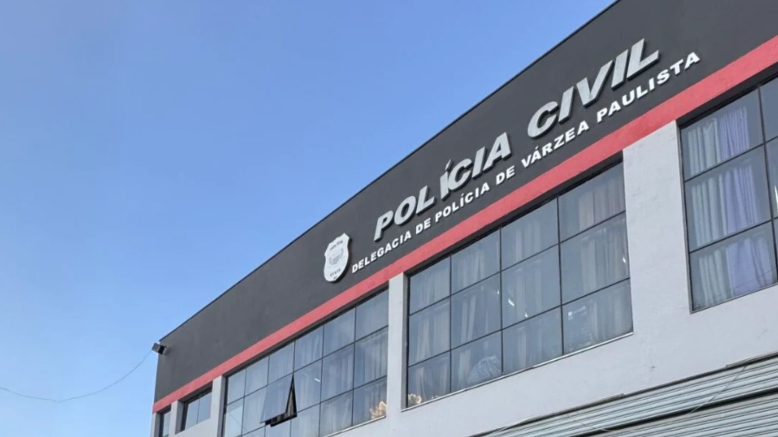 Fachada da Delegacia de Polícia Civil de Várzea Paulista vista de baixo, com o letreiro e o céu azul ao fundo.