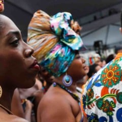 Mulher negra de perfil usando turbante colorido e brinco circular grande, cercada por outras pessoas com roupas e acessórios estampados em um evento cultural.