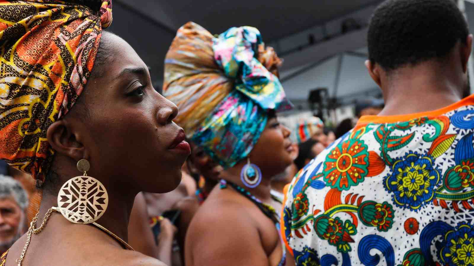 Mulher negra de perfil usando turbante colorido e brinco circular grande, cercada por outras pessoas com roupas e acessórios estampados em um evento cultural.