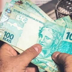 Mãos segurando cédulas de 100 reais, representando pagamento ou movimentação de dinheiro no Brasil.