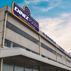 Fachada do prédio industrial da Diniz Foods em Louveira, com letreiro azul e amarelo e céu colorido ao fundo.