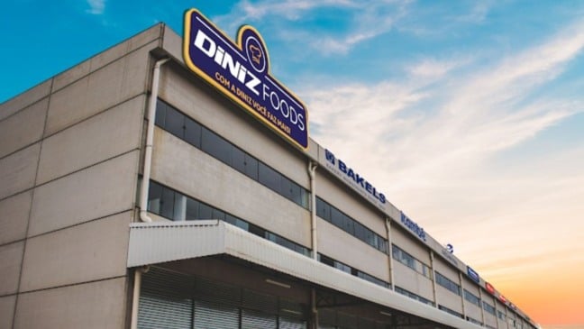 Fachada do prédio industrial da Diniz Foods em Louveira, com letreiro azul e amarelo e céu colorido ao fundo.