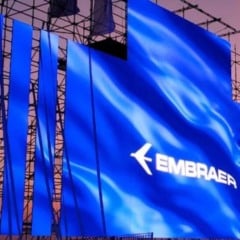 Painel azul com o logo da Embraer montado em uma grande estrutura metálica, iluminado ao pôr do sol durante um evento externo.