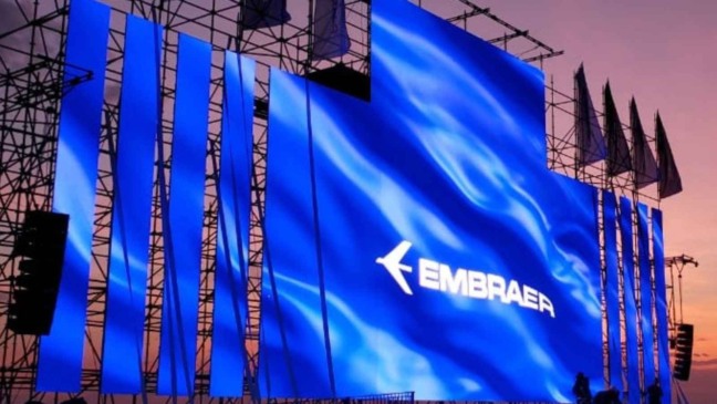 Painel azul com o logo da Embraer montado em uma grande estrutura metálica, iluminado ao pôr do sol durante um evento externo.