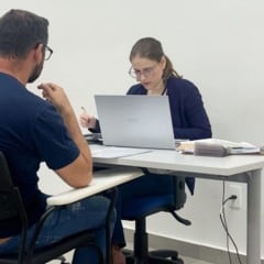 Profissional atendendo um homem sentado, utilizando um notebook e anotando informações em mesa de atendimento, em ambiente claro e organizado.