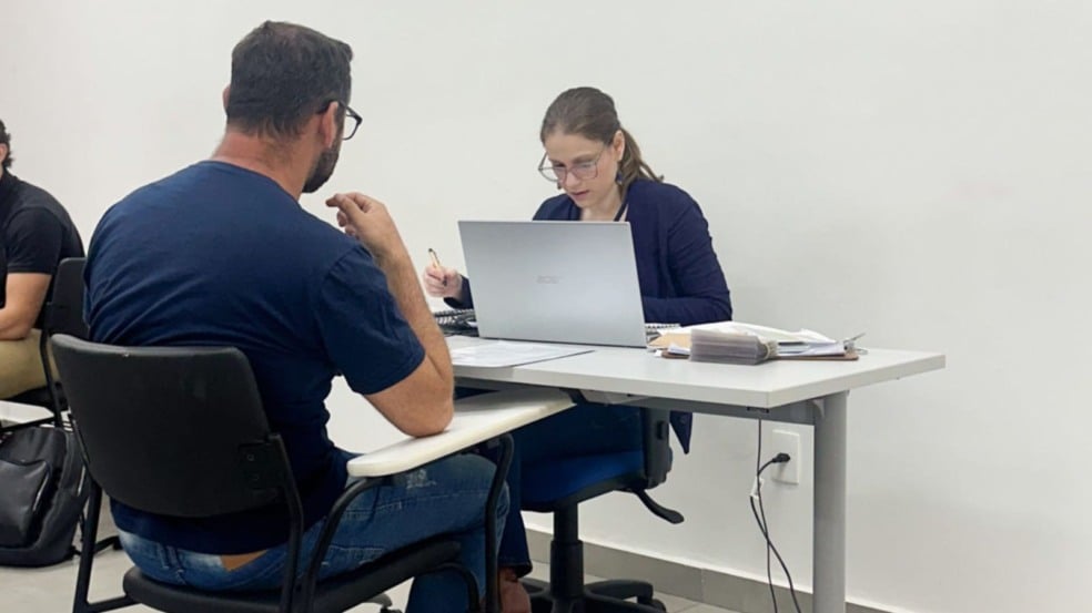 Profissional atendendo um homem sentado, utilizando um notebook e anotando informações em mesa de atendimento, em ambiente claro e organizado. Profissional atendendo um homem sentado, utilizando um notebook e anotando informações em mesa de atendimento, em ambiente claro e organizado.