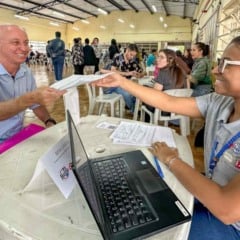 Atendente jovem, sorridente, entrega um documento a um homem em uma mesa com computador durante um evento de emprego em um salão cheio de candidatos.