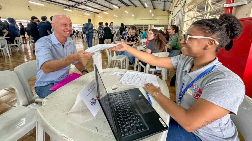 Atendente jovem, sorridente, entrega um documento a um homem em uma mesa com computador durante um evento de emprego em um salão cheio de candidatos.
