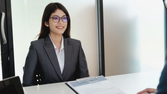 Mulher jovem sorrindo durante entrevista de emprego, usando terno cinza e camisa branca, com currículo sobre a mesa.