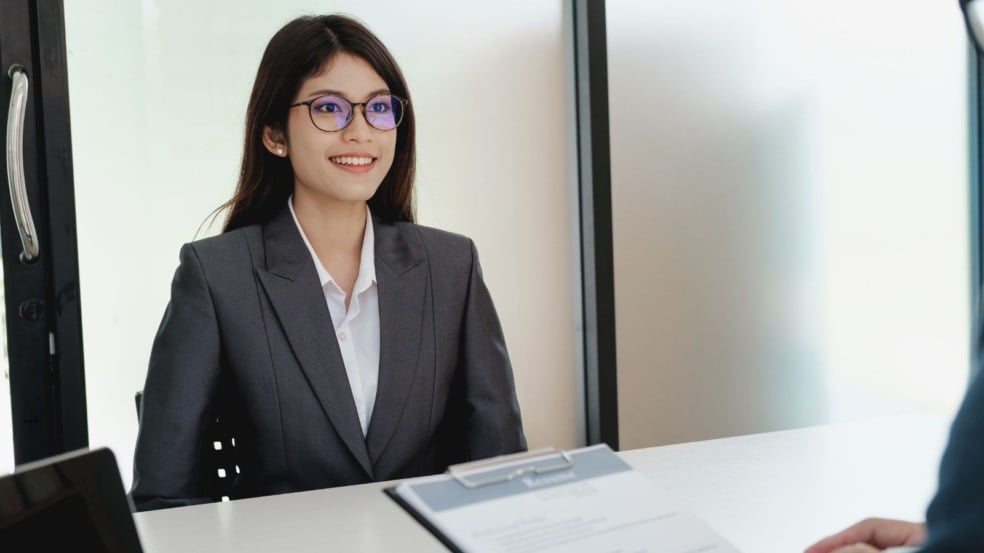Mulher jovem sorrindo durante entrevista de emprego, usando terno cinza e camisa branca, com currículo sobre a mesa.