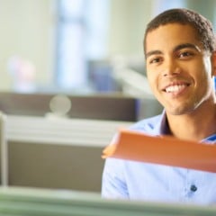 Homem jovem usando camisa azul clara sorri enquanto segura uma pasta alaranjada em um escritório iluminado.