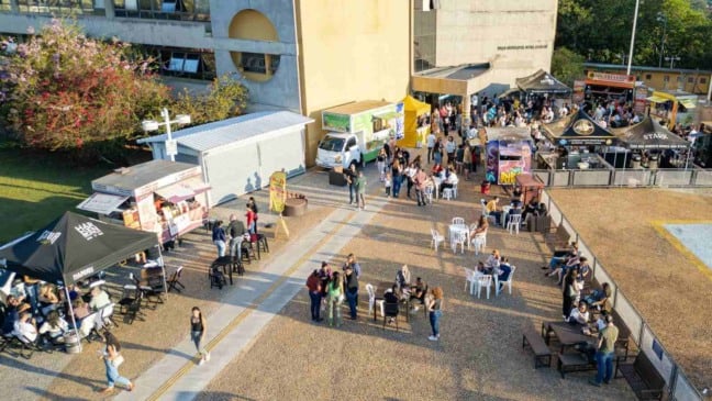Vista aérea do evento Sexta no Paço em Jundiaí, com food trucks, tendas gastronômicas, mesas ocupadas pelo público e a fachada do Paço Municipal ao fundo.
