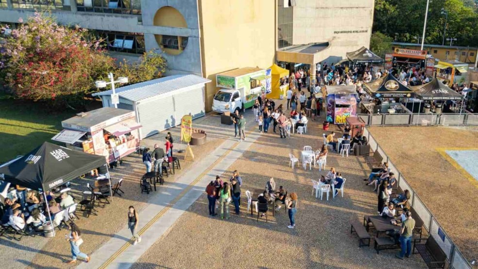 Vista aérea do evento Sexta no Paço em Jundiaí, com food trucks, tendas gastronômicas, mesas ocupadas pelo público e a fachada do Paço Municipal ao fundo.