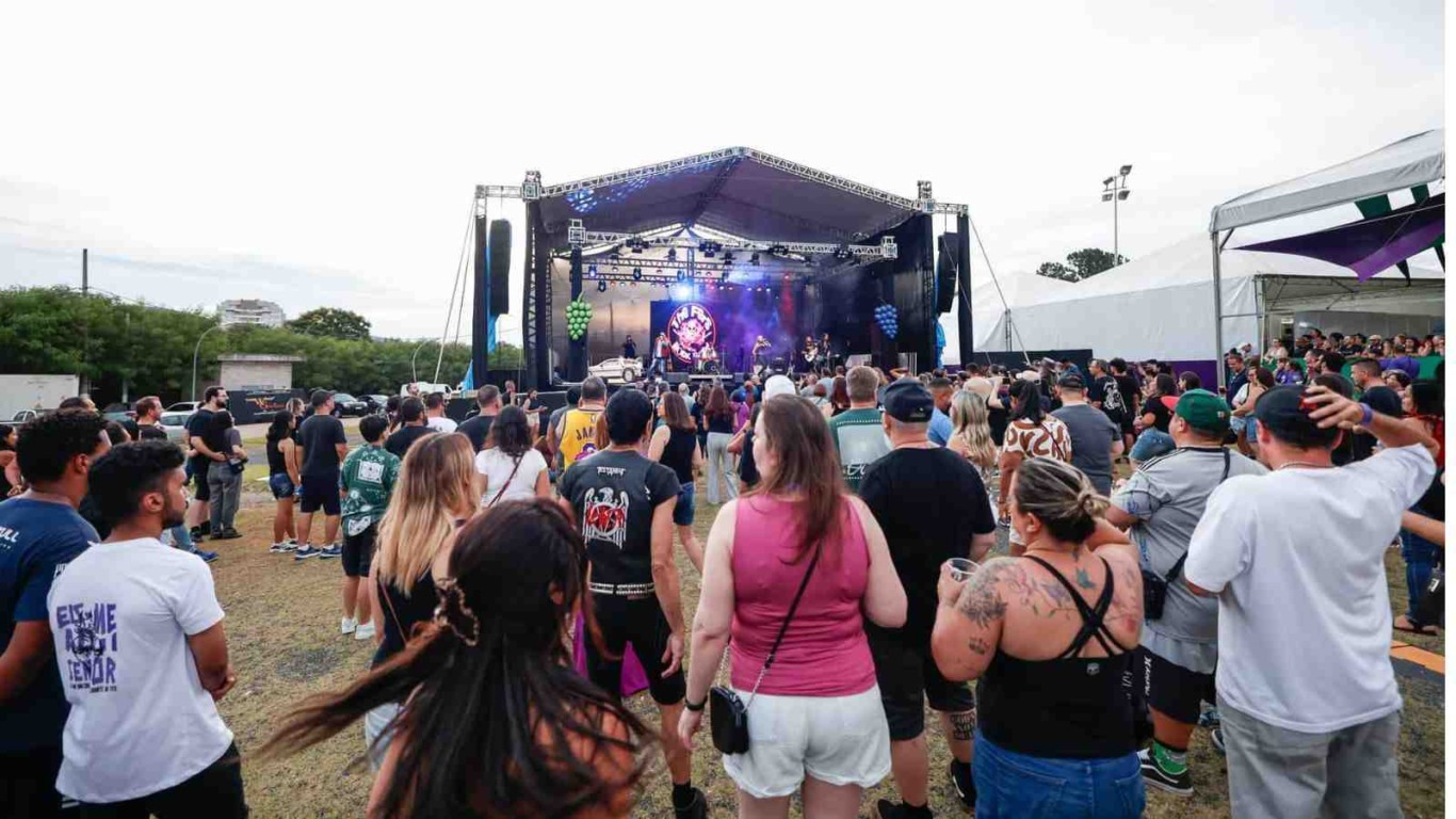 Pessoas reunidas ao ar livre assistindo a um show em um palco grande, com luzes e telão, durante um festival. Pessoas reunidas ao ar livre assistindo a um show em um palco grande, com luzes e telão, durante um festival.