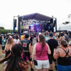 Pessoas reunidas ao ar livre assistindo a um show em um palco grande, com luzes e telão, durante um festival.
