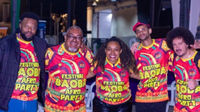 Grupo de cinco pessoas vestindo camisetas coloridas do Festival Baobá Afro Party, sorrindo e celebrando a cultura afro-brasileira.