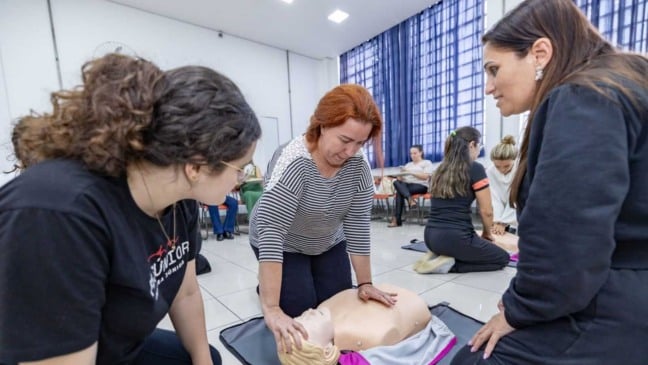 Gestora escolar realiza manobra de RCP em boneco durante treinamento de primeiros socorros em Jundiaí, com orientação de estudantes da FMJúnior.