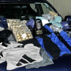 Itens furtados expostos no capô de uma viatura da Guarda Municipal de Jundiaí, incluindo camisetas, bermudas, bonés e bandejas de chocolate, ao lado de um equipamento com o brasão da GMJ.