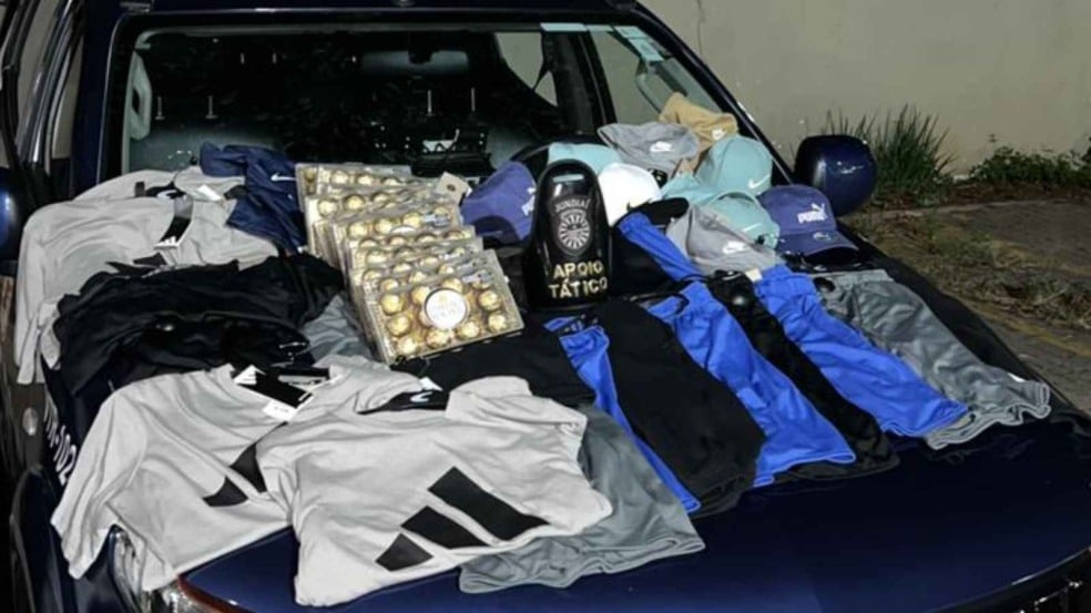 Itens furtados expostos no capô de uma viatura da Guarda Municipal de Jundiaí, incluindo camisetas, bermudas, bonés e bandejas de chocolate, ao lado de um equipamento com o brasão da GMJ. Itens furtados expostos no capô de uma viatura da Guarda Municipal de Jundiaí, incluindo camisetas, bermudas, bonés e bandejas de chocolate, ao lado de um equipamento com o brasão da GMJ.