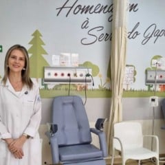 Profissional da saúde de jaleco branco em uma sala pediátrica com poltronas de tratamento e parede decorada com desenhos de árvores e nuvens.