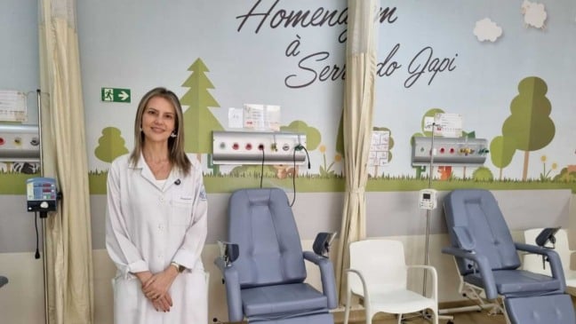 Profissional da saúde de jaleco branco em uma sala pediátrica com poltronas de tratamento e parede decorada com desenhos de árvores e nuvens.