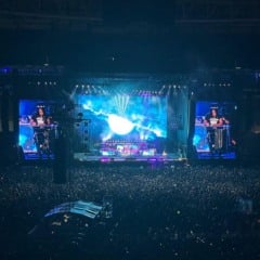 Show do Guns N’ Roses com Axl Rose cantando no telão e Slash tocando guitarra, diante de um estádio lotado iluminado em vermelho.