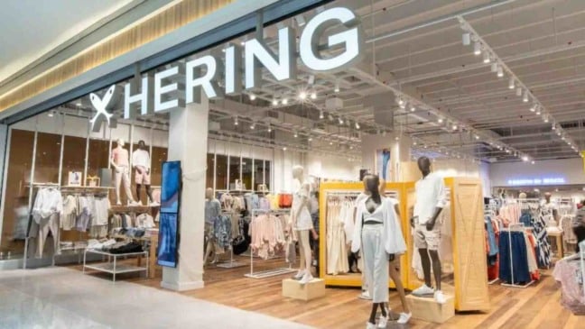 Fachada iluminada de uma loja da Hering em shopping, com manequins e araras de roupas no interior.