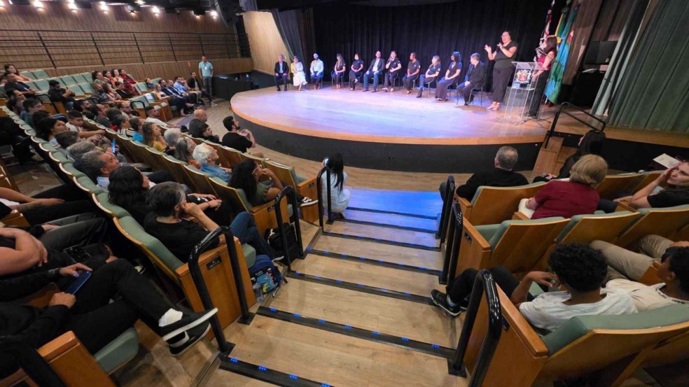 Auditório cheio na Sala Glória Rocha, com público assistindo ao lançamento do Hino de Jundiaí em Libras. No palco, convidados estão sentados em fileira e uma intérprete de Libras atua ao lado do púlpito.