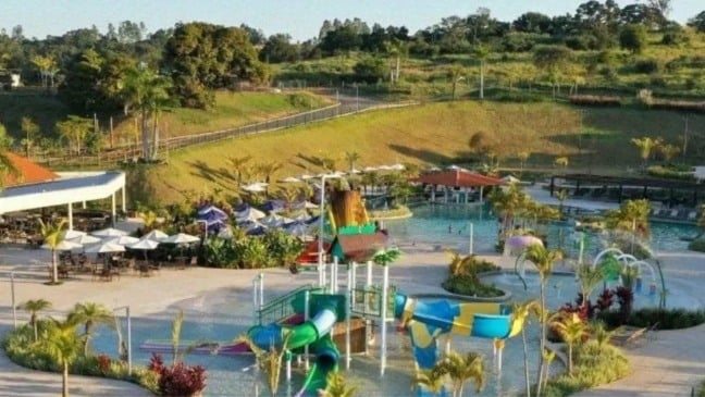 Vista de um parque aquático com piscinas, escorregadores infantis, guarda-sóis e áreas verdes ao redor, em ambiente de resort.