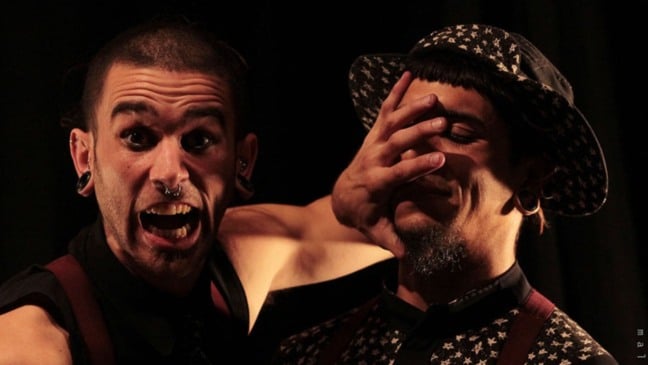 Dois artistas de circo em cena; um deles faz expressão intensa com a boca aberta enquanto cobre o rosto do outro com a mão, criando efeito cômico e teatral.