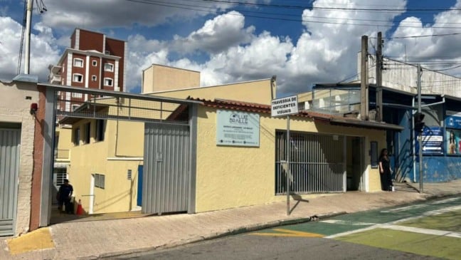 Fachada do Instituto Luiz Braille em Jundiaí, com prédio amarelo, portão cinza, placa de identificação e placa de “Travessia de Deficientes Visuais” em frente ao local.