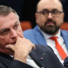 “Homem de terno escuro sentado com expressão pensativa e mão no queixo, enquanto outros homens de terno aparecem ao fundo em um ambiente formal.”