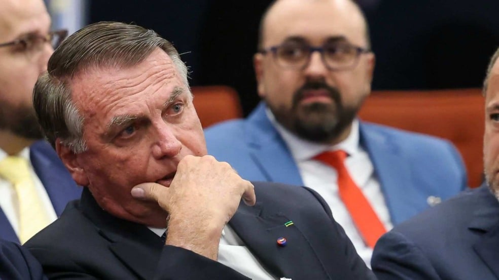 “Homem de terno escuro sentado com expressão pensativa e mão no queixo, enquanto outros homens de terno aparecem ao fundo em um ambiente formal.” “Homem de terno escuro sentado com expressão pensativa e mão no queixo, enquanto outros homens de terno aparecem ao fundo em um ambiente formal.”