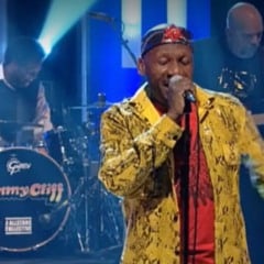 Jimmy Cliff cantando ao vivo no palco, vestindo jaqueta amarela estampada e segurando um microfone, com banda e bateria ao fundo exibindo seu nome.