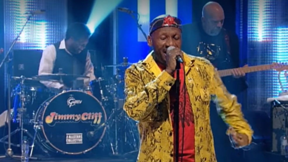 Jimmy Cliff cantando ao vivo no palco, vestindo jaqueta amarela estampada e segurando um microfone, com banda e bateria ao fundo exibindo seu nome. Jimmy Cliff cantando ao vivo no palco, vestindo jaqueta amarela estampada e segurando um microfone, com banda e bateria ao fundo exibindo seu nome.