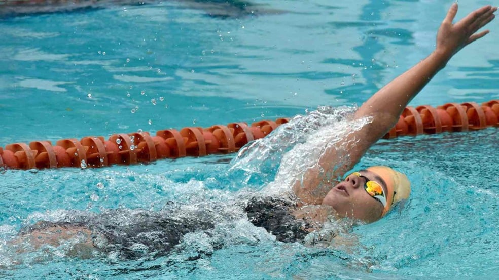 Nadadora praticando nado costas em piscina, com touca clara, óculos espelhados e raia laranja ao fundo. Nadadora praticando nado costas em piscina, com touca clara, óculos espelhados e raia laranja ao fundo.