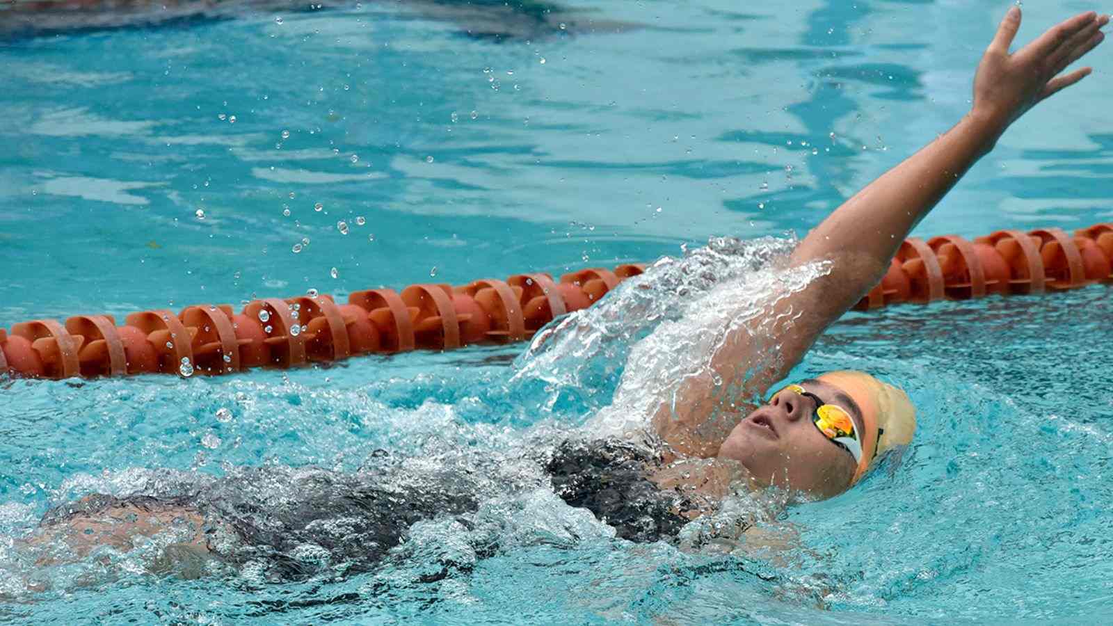 Nadadora praticando nado costas em piscina, com touca clara, óculos espelhados e raia laranja ao fundo.