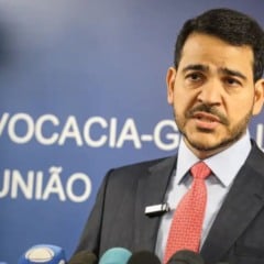 Homem de terno e gravata vermelha fala à imprensa diante de microfones, com painel azul da Advocacia-Geral da União ao fundo.