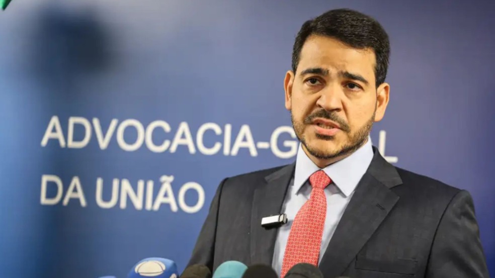 Homem de terno e gravata vermelha fala à imprensa diante de microfones, com painel azul da Advocacia-Geral da União ao fundo. Homem de terno e gravata vermelha fala à imprensa diante de microfones, com painel azul da Advocacia-Geral da União ao fundo.