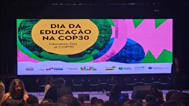 Telão do “Dia da Educação na COP30” em um auditório, com pessoas circulando à frente do palco antes do início das atividades.
