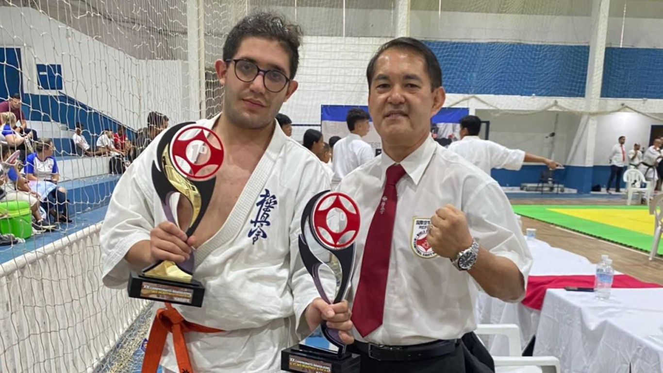 Atleta e sensei posando com troféus e punhos erguidos após conquista no Campeonato Brasileiro de Karatê.