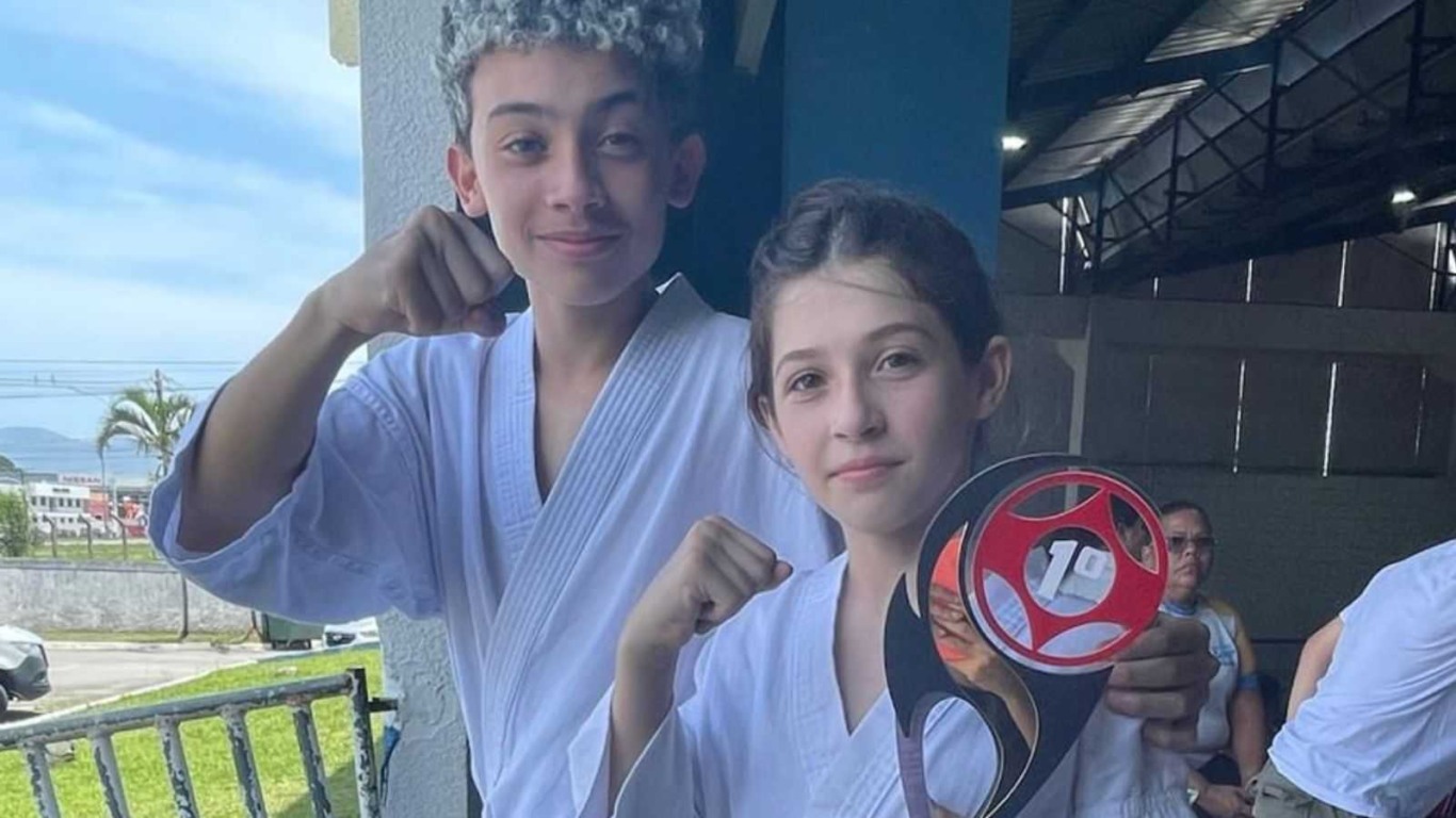 Dois jovens karatecas de Jundiaí, um segurando o troféu de primeiro lugar, posam com os punhos erguidos após a vitória no Campeonato Brasileiro.