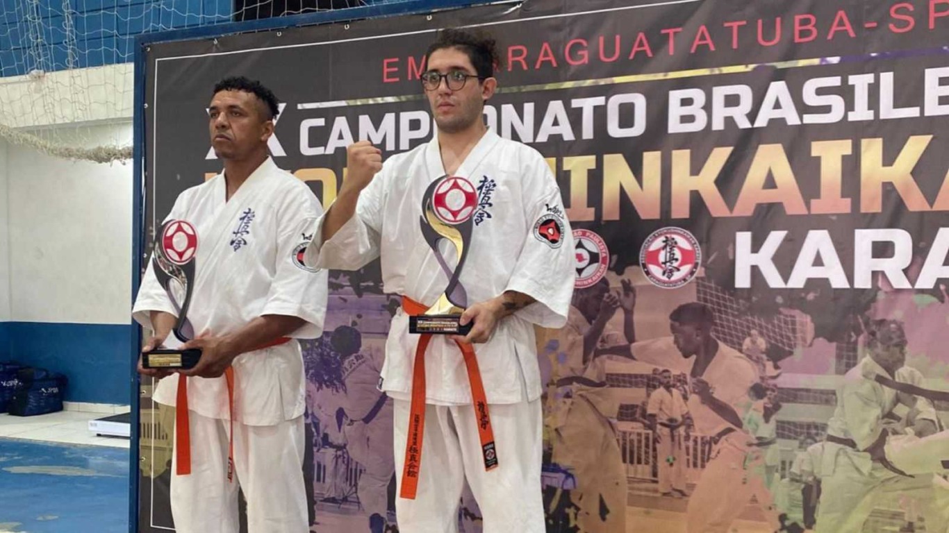 Dois atletas de karatê de Jundiaí, de quimono branco e faixa laranja, seguram troféus em frente ao painel do Campeonato Brasileiro.