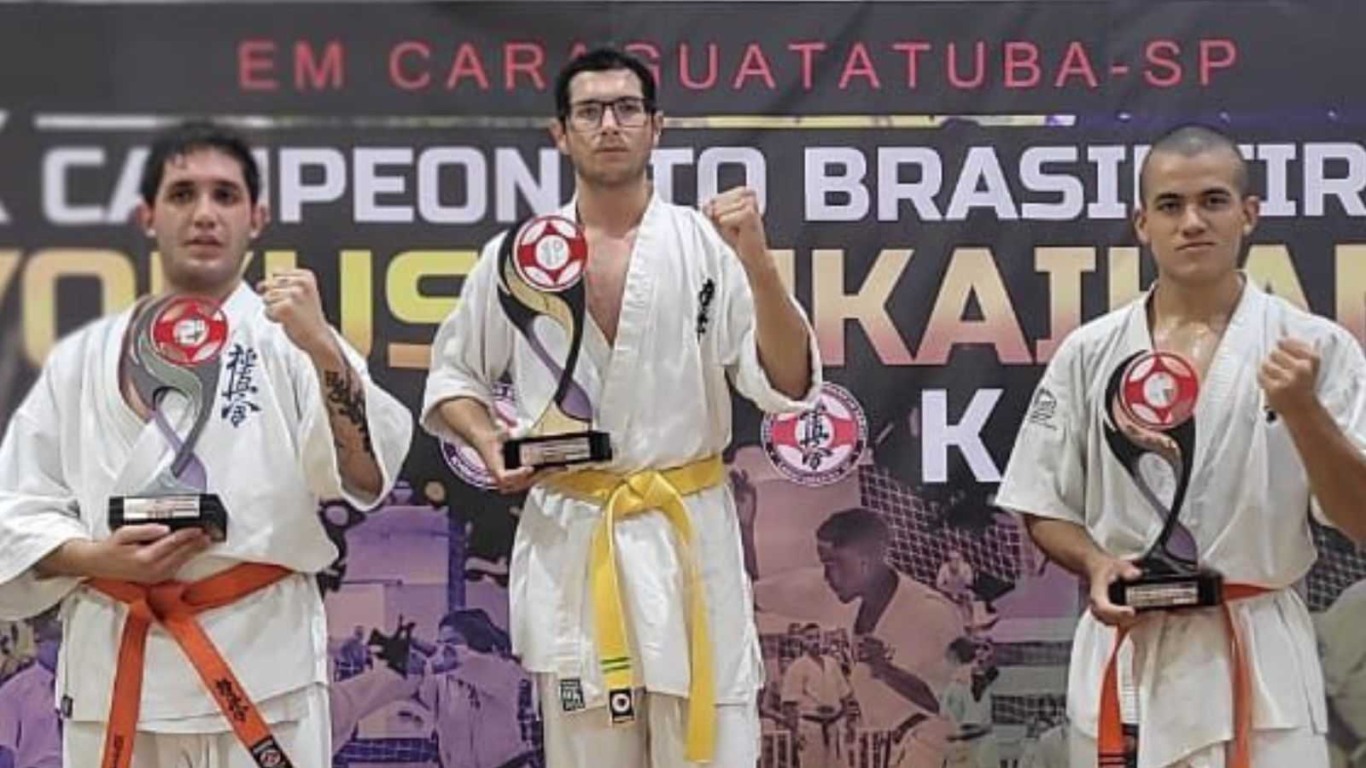 Três atletas de karatê de Jundiaí segurando troféus e fazendo pose de luta em frente ao banner do Campeonato Brasileiro em Caraguatatuba.