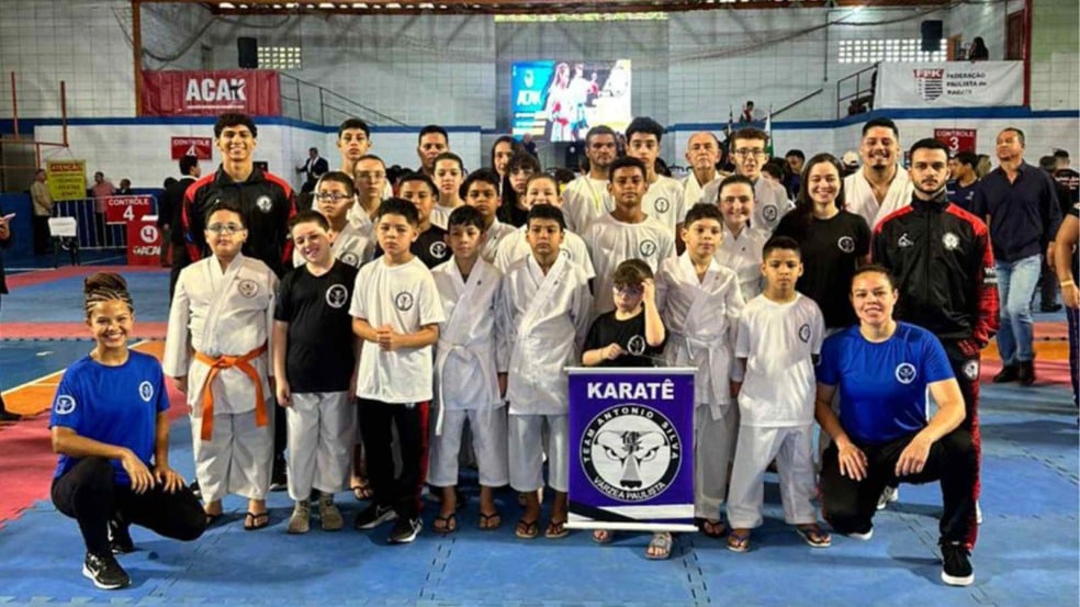 Equipe de karatê de Várzea Paulista reunida no tatame para foto em grupo durante a 9ª Copa de Karatê, segurando estandarte da equipe Team Antonio Silva.