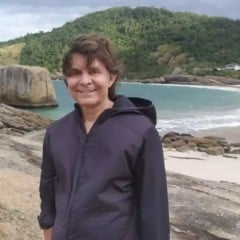 Lô Borges sorrindo em uma praia cercada por montanhas e mar, usando jaqueta escura sobre as rochas.
