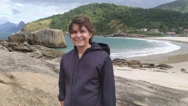 Lô Borges sorrindo em uma praia cercada por montanhas e mar, usando jaqueta escura sobre as rochas.