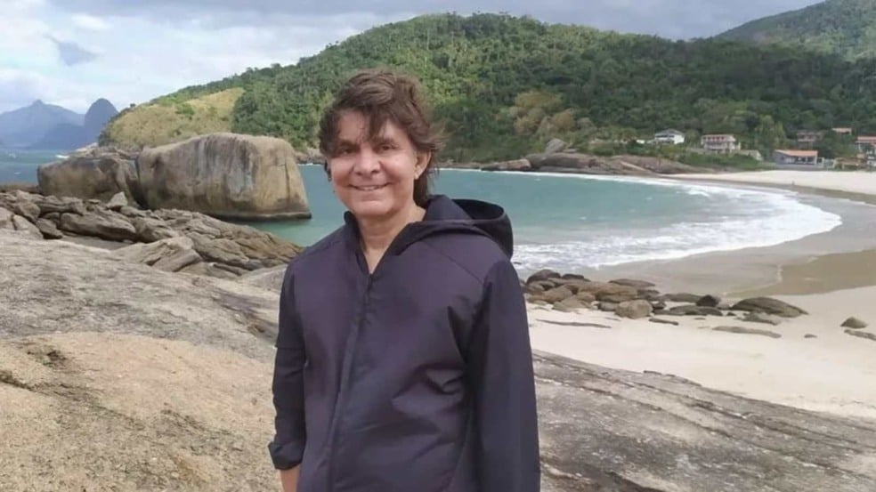 Lô Borges sorrindo em uma praia cercada por montanhas e mar, usando jaqueta escura sobre as rochas.