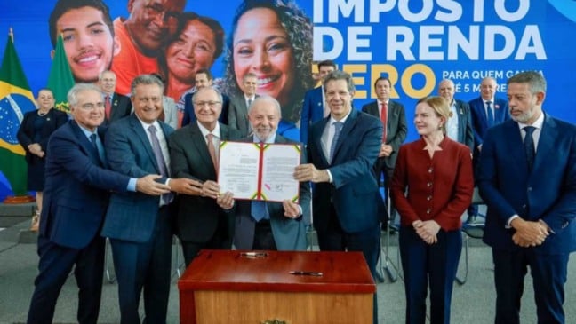Presidente Lula, ao centro, segurando o documento de sanção da nova lei do Imposto de Renda ao lado de ministros e autoridades, diante de painel azul com a frase “Imposto de Renda Zero”.